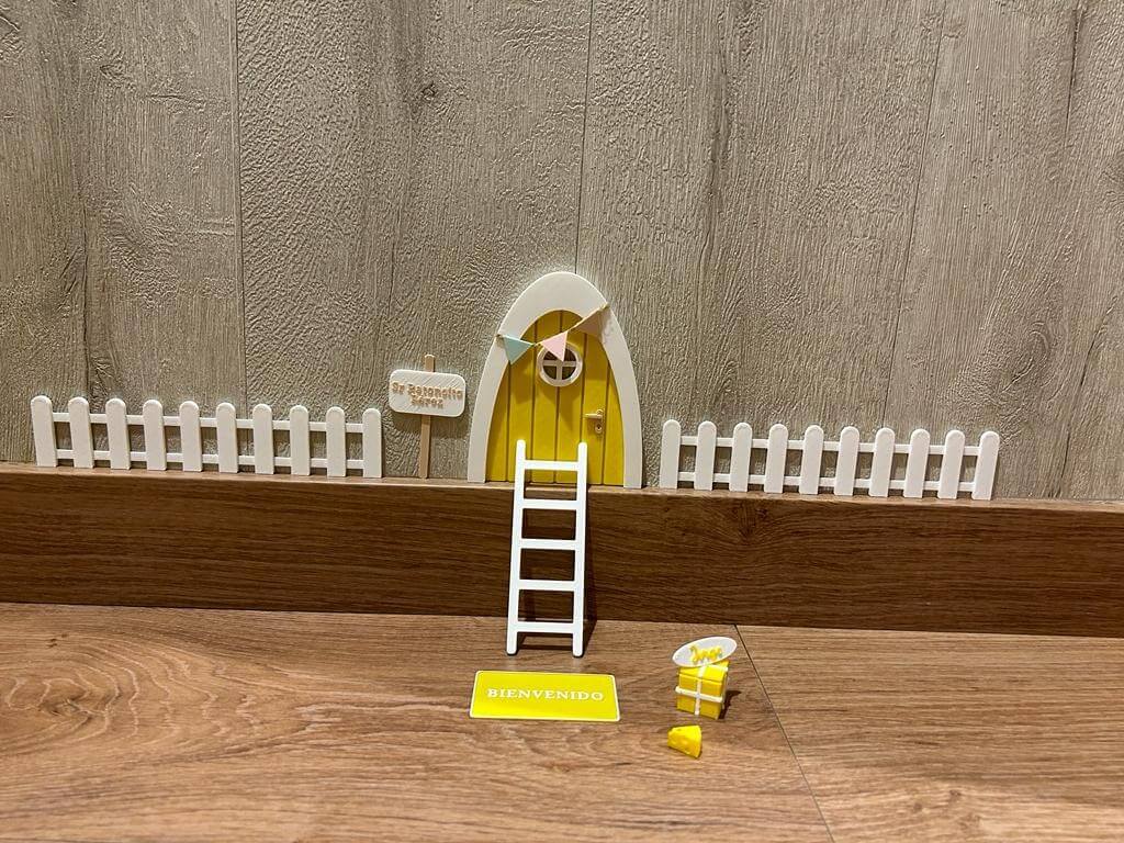 Puerta Ratón Pérez con puerta en miniatura amarillo, escalera, valla, cartel de bienvenida y queso, creando un escenario encantador en 3D sobre una superficie de madera.