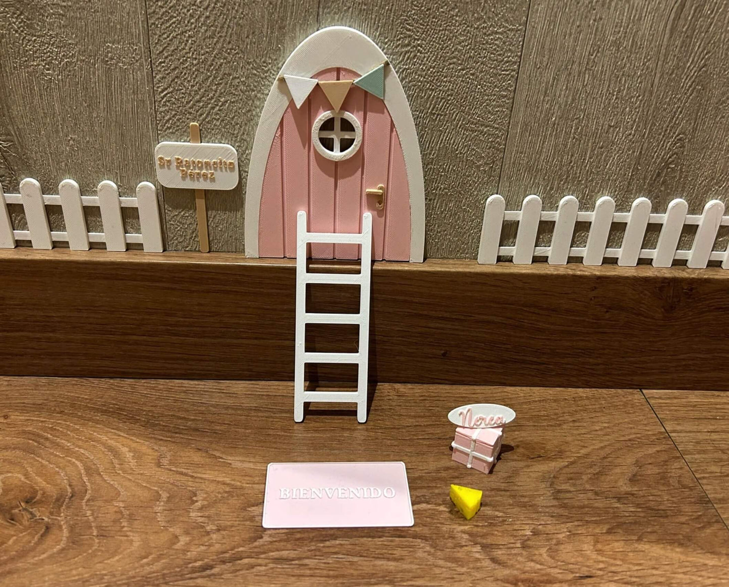 Puerta del Ratoncito Pérez, puerta en rosa pastel, escalera, valla, cartel de bienvenida y queso, creando un escenario encantador en 3D sobre una superficie de madera.