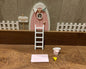 Puerta del Ratoncito Pérez, puerta en rosa pastel, escalera, valla, cartel de bienvenida y queso, creando un escenario encantador en 3D sobre una superficie de madera.